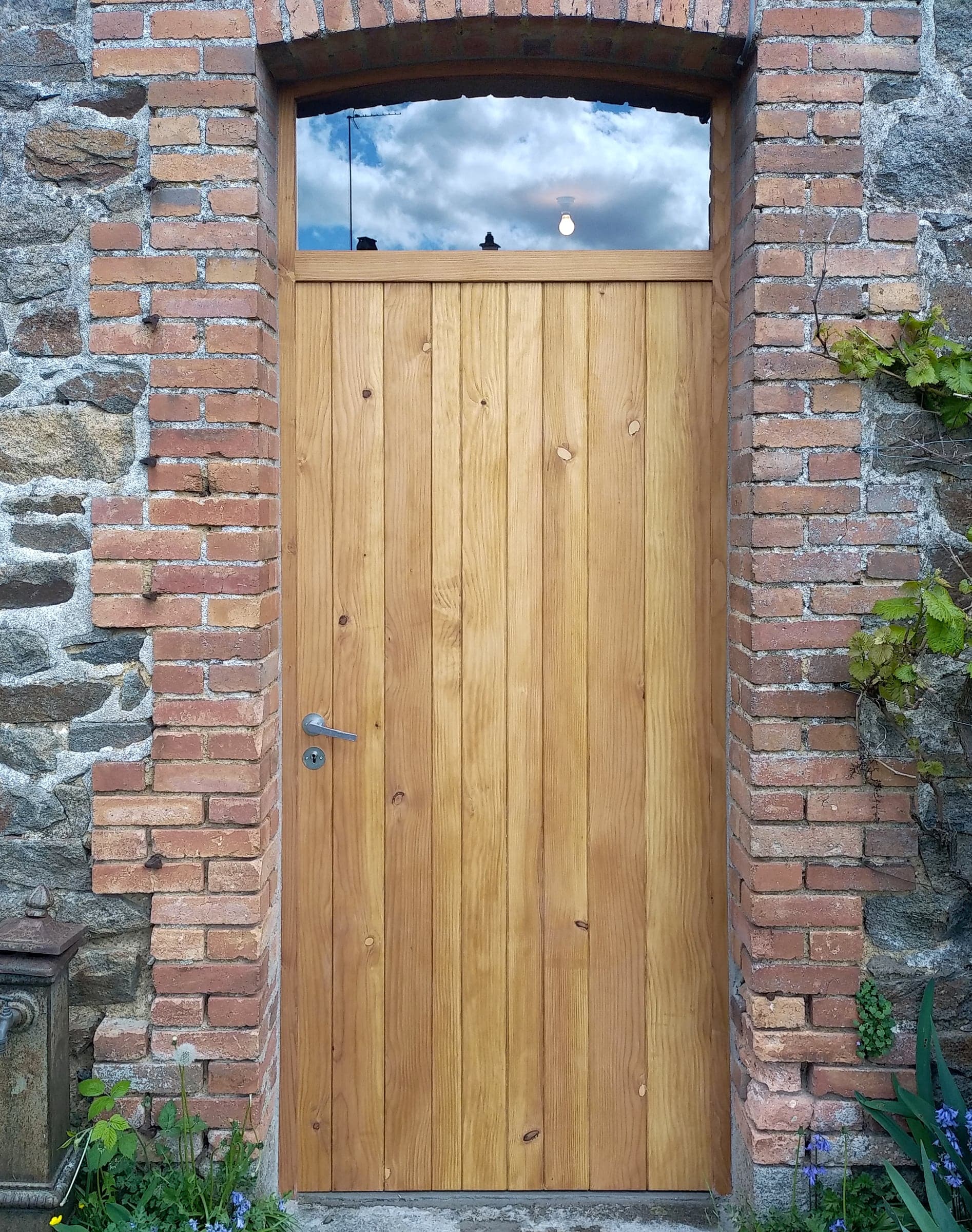 Porte en bois massif à lames verticales avec imposte vitrée, sous un arc de brique